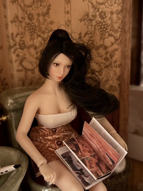 Olive Joanne, a miniature doll with long dark hair, reading a tiny magazine in front of patterned wallpaper